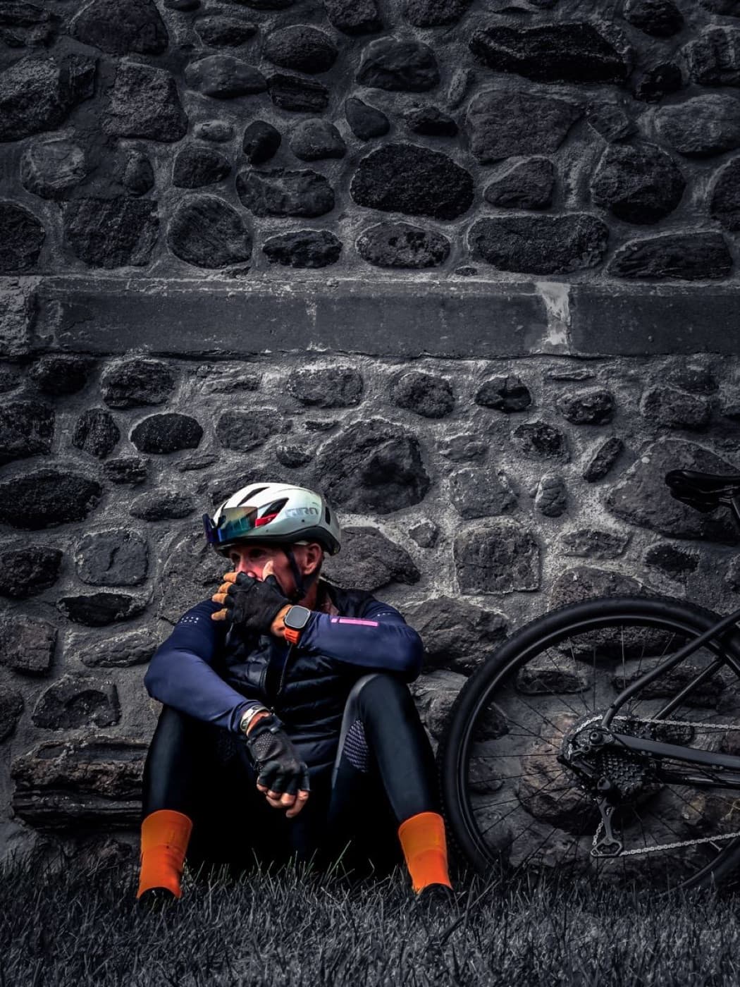 Ciclista sentado y agotado, apoyado contra un muro de piedra junto a su bici